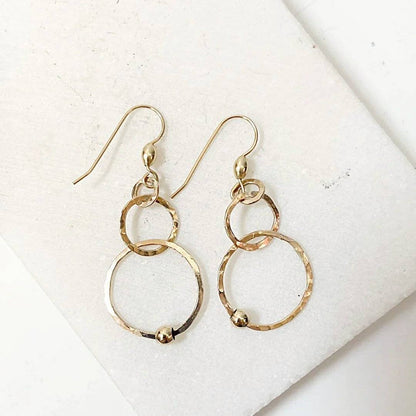 Pair of gold HoopLink Earrings with interlinked hammered hoops and bead detail, laid flat on a white stone surface for product display.