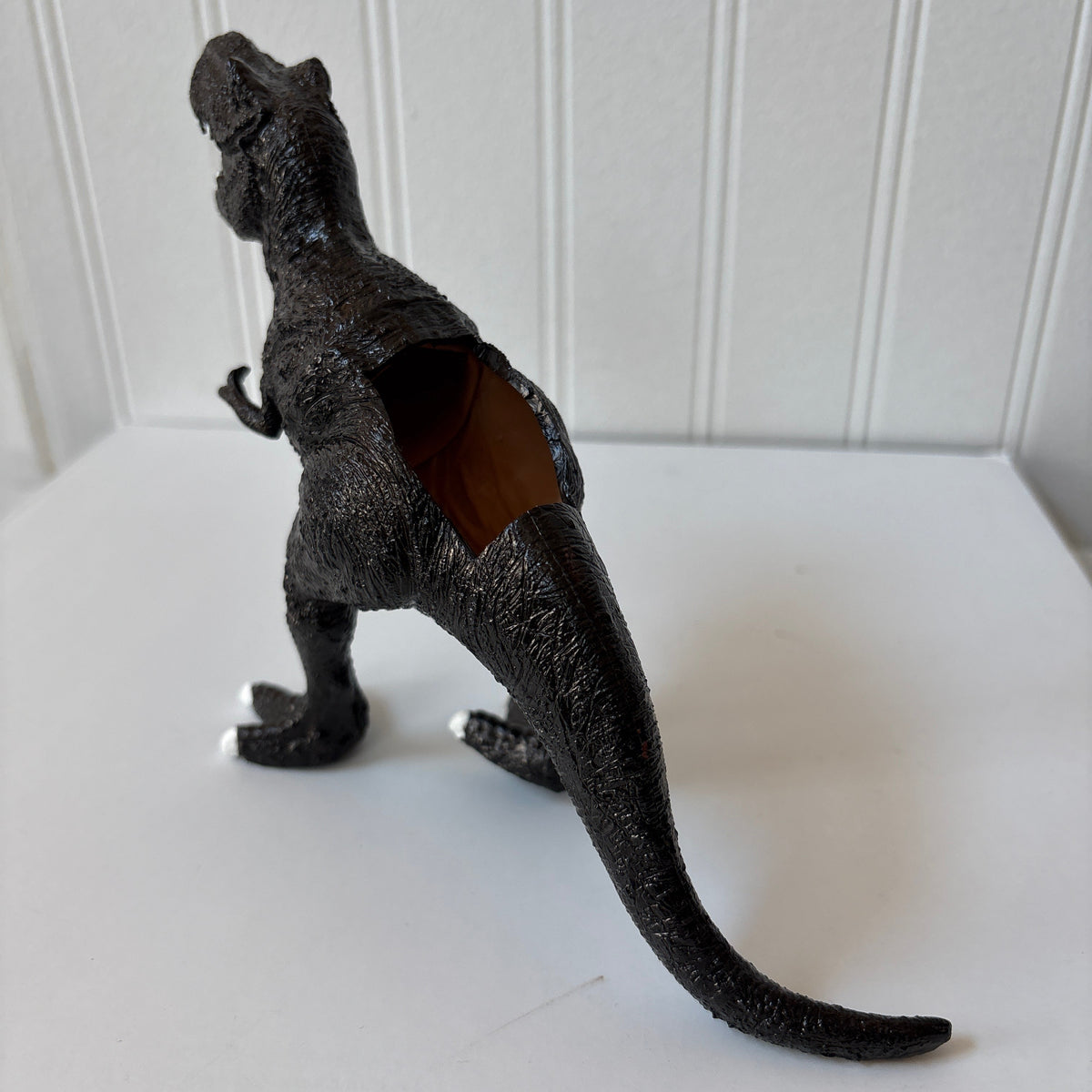 A black textured dinosaur figurine, shaped like a T-rex with a hollowed-out body revealing a brown interior, standing on a white surface against a white paneled wall.