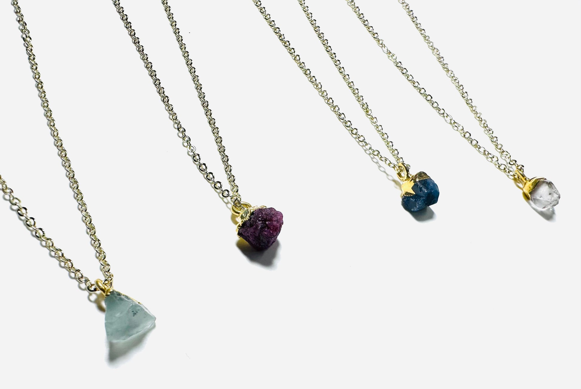 Four raw gemstone necklaces featuring aquamarine, ruby, sapphire, and Herkimer diamond on gold brass chains. UNI-T Jewelry gift collection.