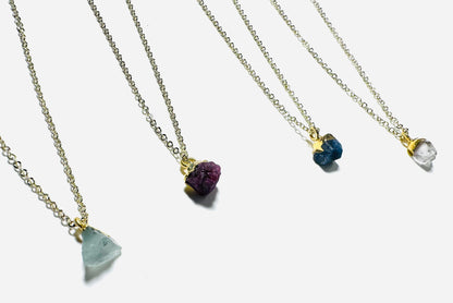 Four raw gemstone necklaces featuring aquamarine, ruby, sapphire, and Herkimer diamond on gold brass chains. UNI-T Jewelry gift collection.
