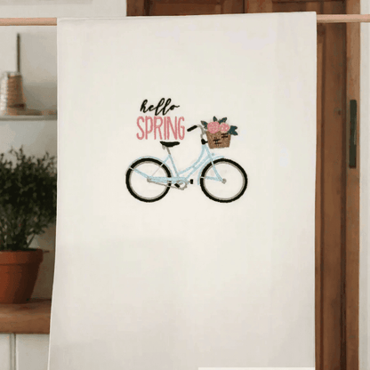 Spring Bike Embroidered Towel hanging on a wooden cabinet, showcasing a detailed blue bike and 'Hello Spring' text for seasonal kitchen decor.