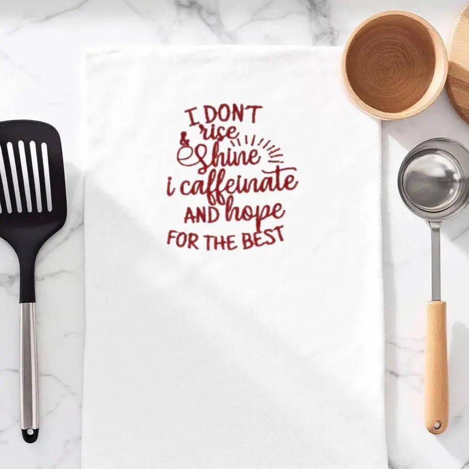 Embroidered towel with coffee humor quote 'I don't rise and shine, I caffeinate and hope for the best' in red, ideal kitchen accessory for coffee lovers.
