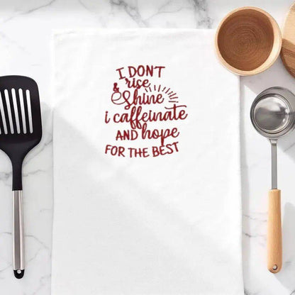 Embroidered towel with coffee humor quote 'I don't rise and shine, I caffeinate and hope for the best' in red, ideal kitchen accessory for coffee lovers.