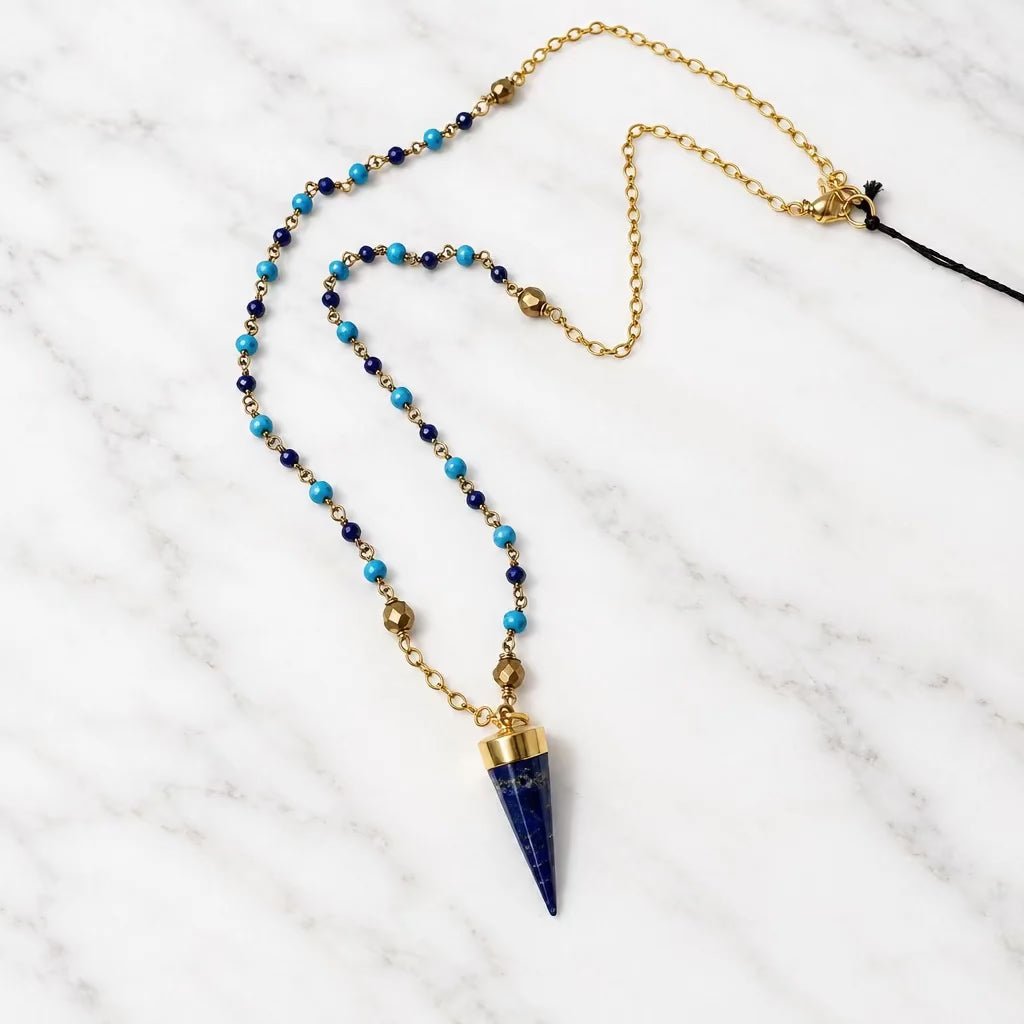 A gold chain necklace featuring a cone-shaped dark blue lapis lazuli pendant, accented with small turquoise and dark brown beads, laid out on a white surface.