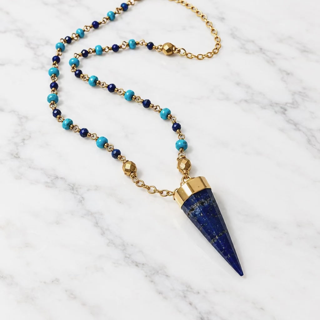 A beaded necklace featuring a dark blue lapis lazuli cone pendant, gold-toned metal chain and caps, and small turquoise and dark blue beads, displayed on a light off-white surface.