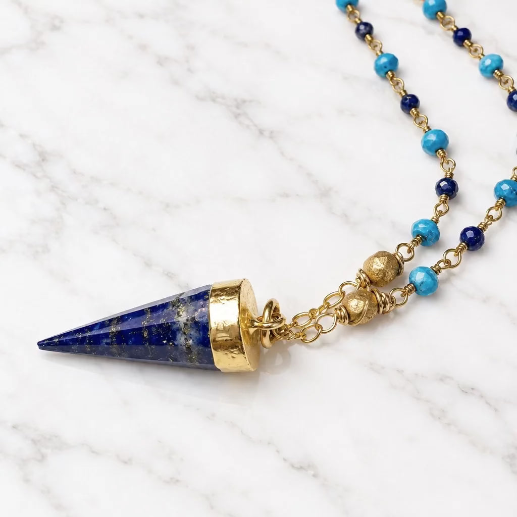 A gold-toned beaded necklace featuring a pointed lapis lazuli pendant and turquoise-blue beads, displayed on a light off-white surface.