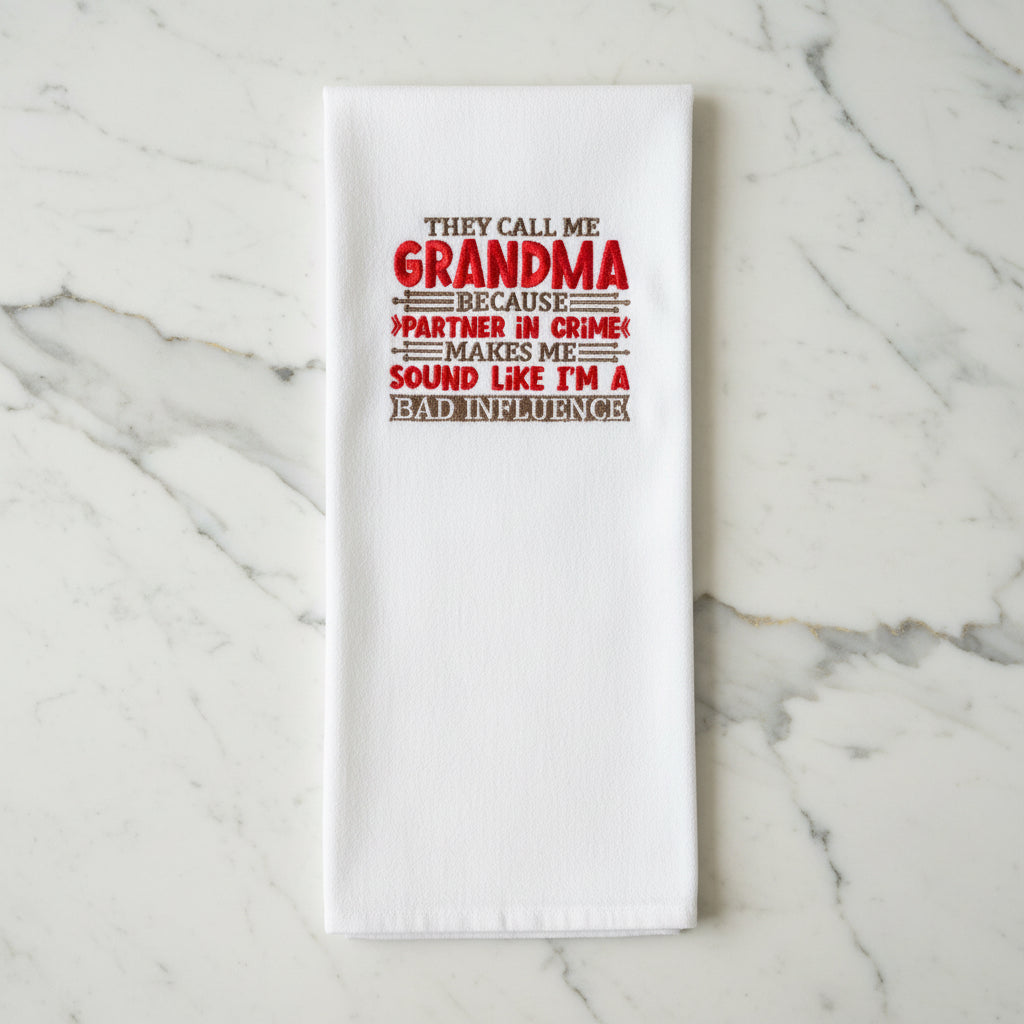 A white kitchen towel hanging in a clear display case, printed with a humorous quote about being called 'Grandma' because it makes you sound like 'I'm a bad influence' in red and brown lettering.