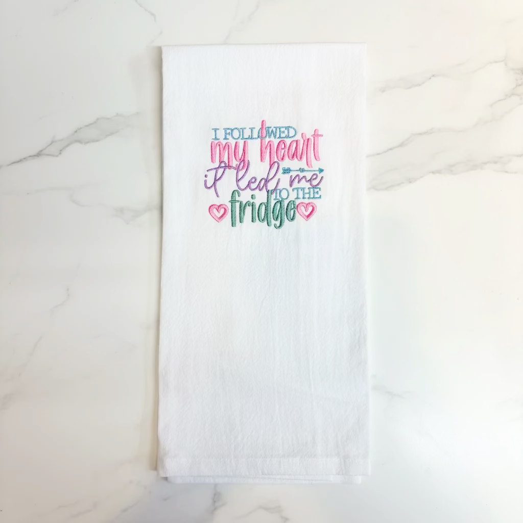 A white kitchen towel hanging from a black plastic hanger against a plain off-white wall. The towel features colorful embroidered text that reads 'I FOLLOWED my heart it led me TO THE fridge' in pink, blue, and green lettering with small heart details.