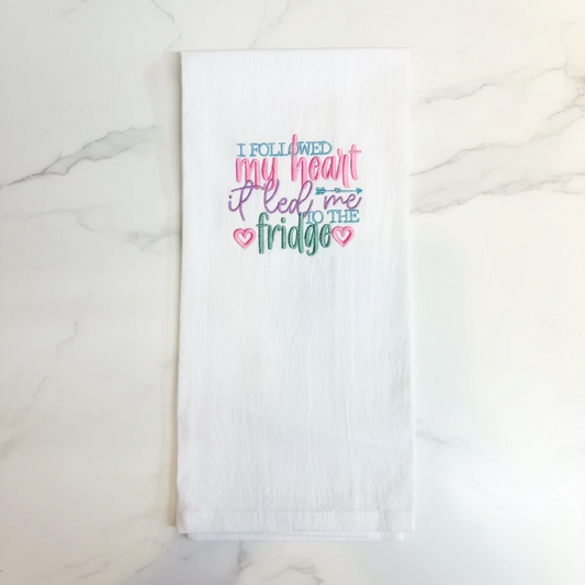 A white kitchen towel hanging from a black plastic hanger against a plain off-white wall. The towel features colorful embroidered text that reads 'I FOLLOWED my heart it led me TO THE fridge' in pink, blue, and green lettering with small heart details.