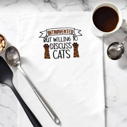 Embroidered towel with 'Introverted but willing to discuss cats' text and paw design, styled on marble countertop with coffee and utensils. Quirky kitchen decor for cat lovers.