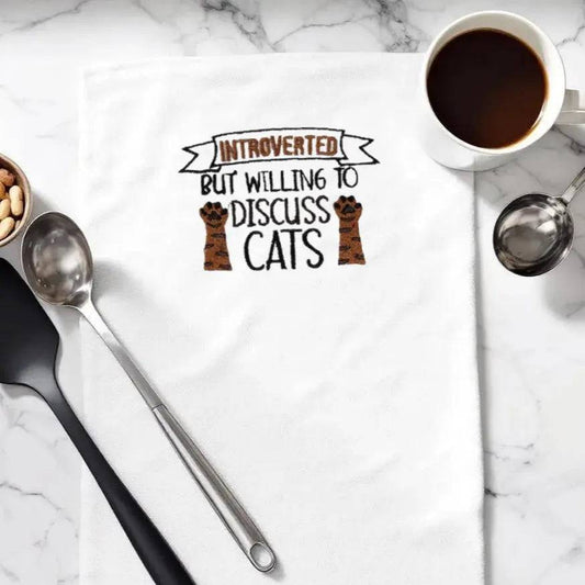 Embroidered towel with 'Introverted but willing to discuss cats' text and paw design, styled on marble countertop with coffee and utensils. Quirky kitchen decor for cat lovers.