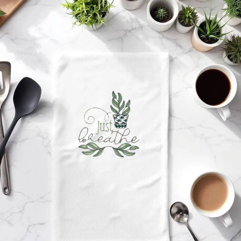 Just Breathe plant towel embroidered with green leaves and potted plant, styled on kitchen counter with coffee and utensils. Ideal kitchen decor gift.