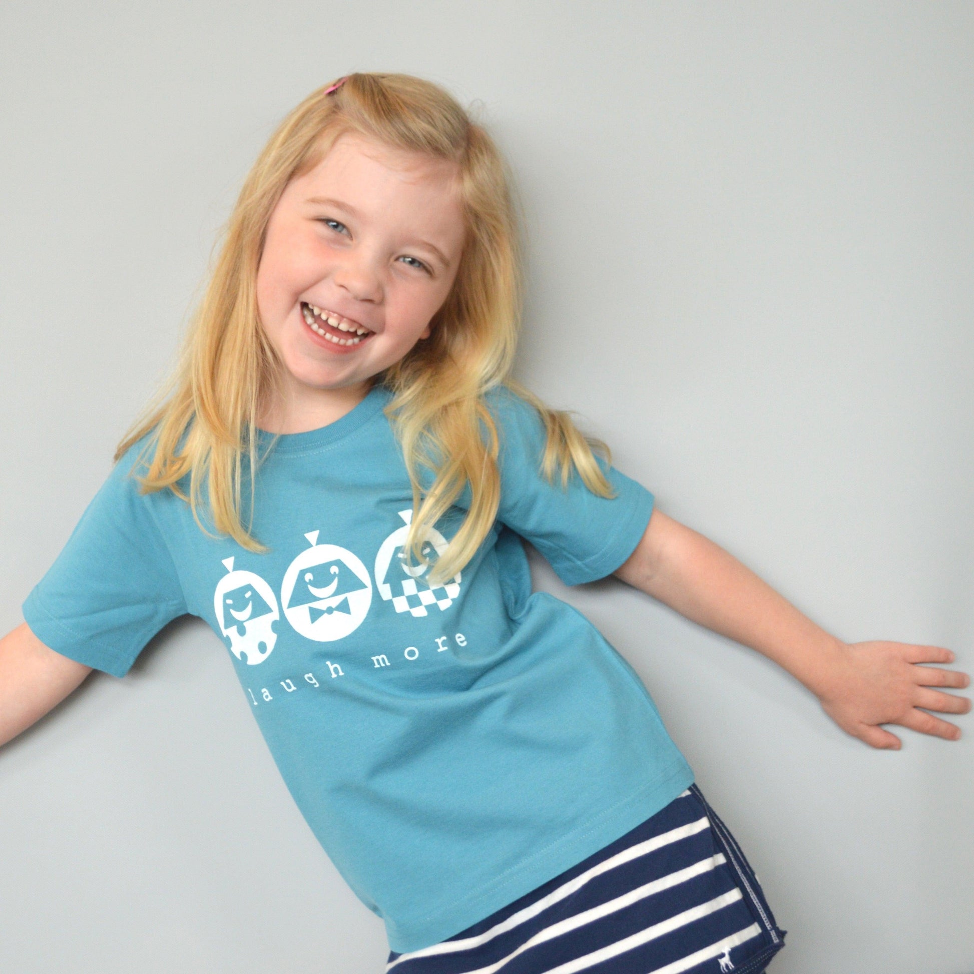 Smiling child wearing Kids' Happy Vibe Organic Cotton T-Shirt in teal blue with 'Laugh More' graphic. Eco-friendly playful shirt for children.