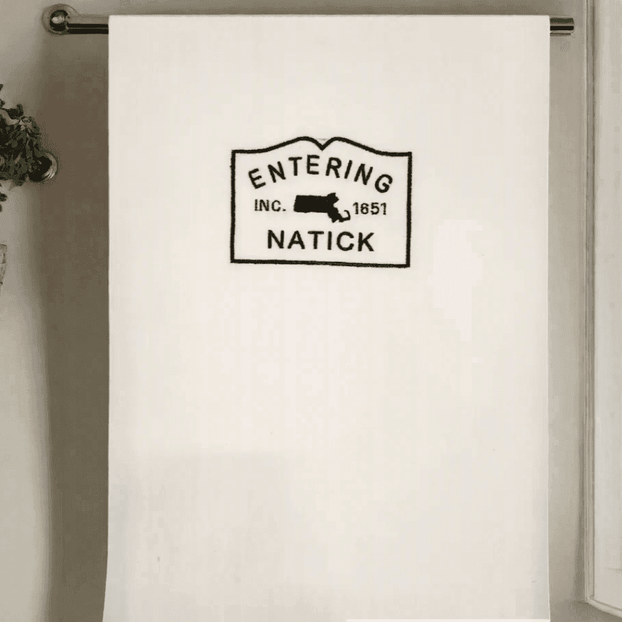 Natick Town Sign Towel hanging on a rod, showcasing black embroidery on white fabric. Perfect kitchen towel for Natick-themed home décor.