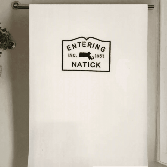 Natick Town Sign Towel hanging on a rod, showcasing black embroidery on white fabric. Perfect kitchen towel for Natick-themed home décor.