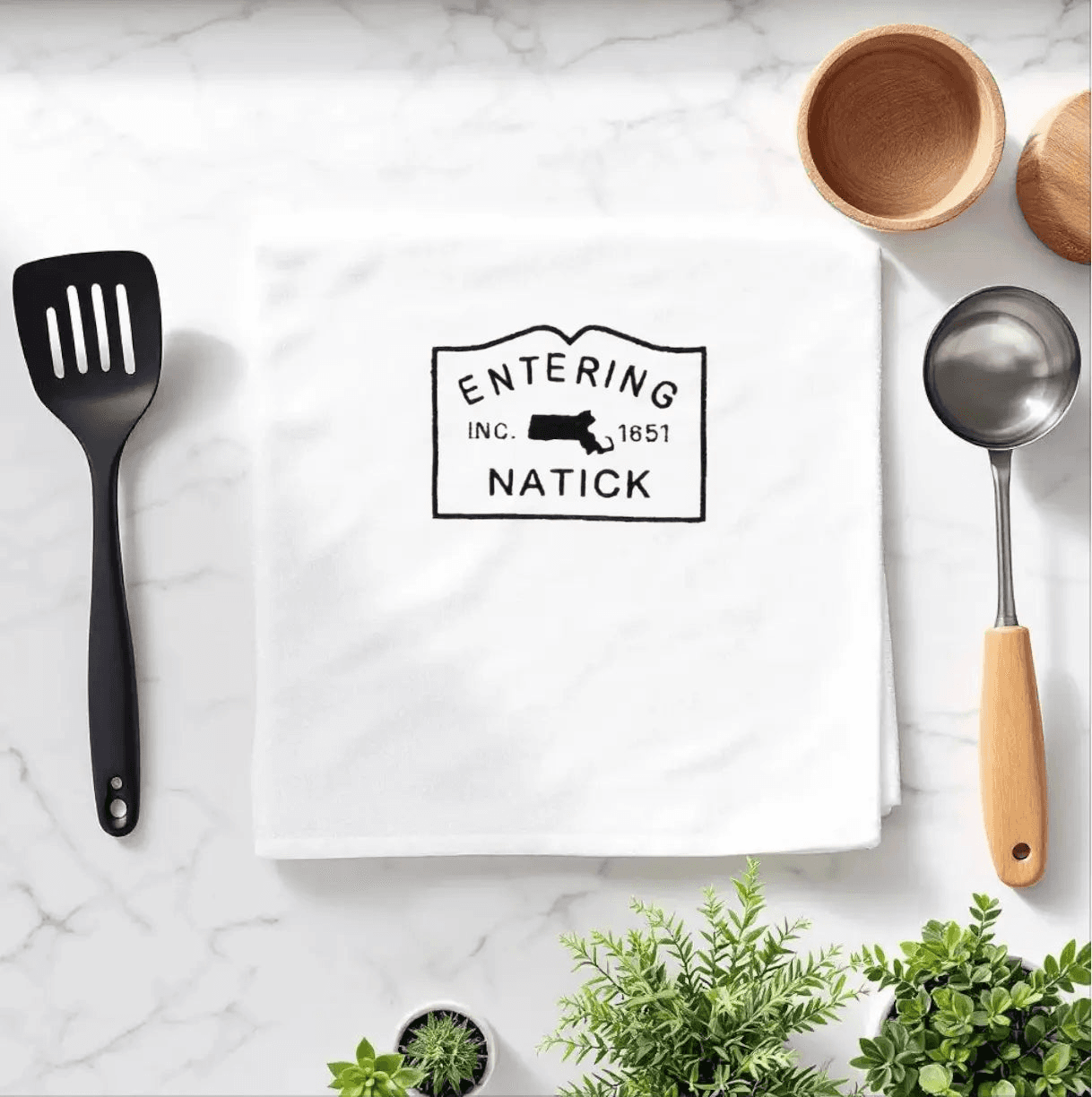 Natick Towel featuring embroidered 'Entering Natick' town sign, displayed on a marble kitchen counter with utensils. Ideal for kitchen decoration or gifting.
