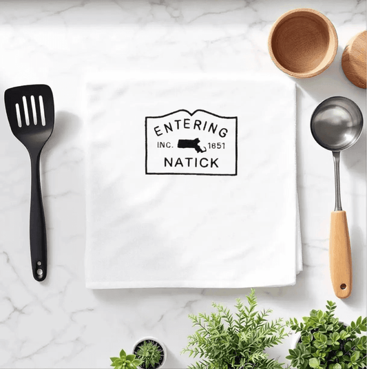 Natick Towel featuring embroidered 'Entering Natick' town sign, displayed on a marble kitchen counter with utensils. Ideal for kitchen decoration or gifting.