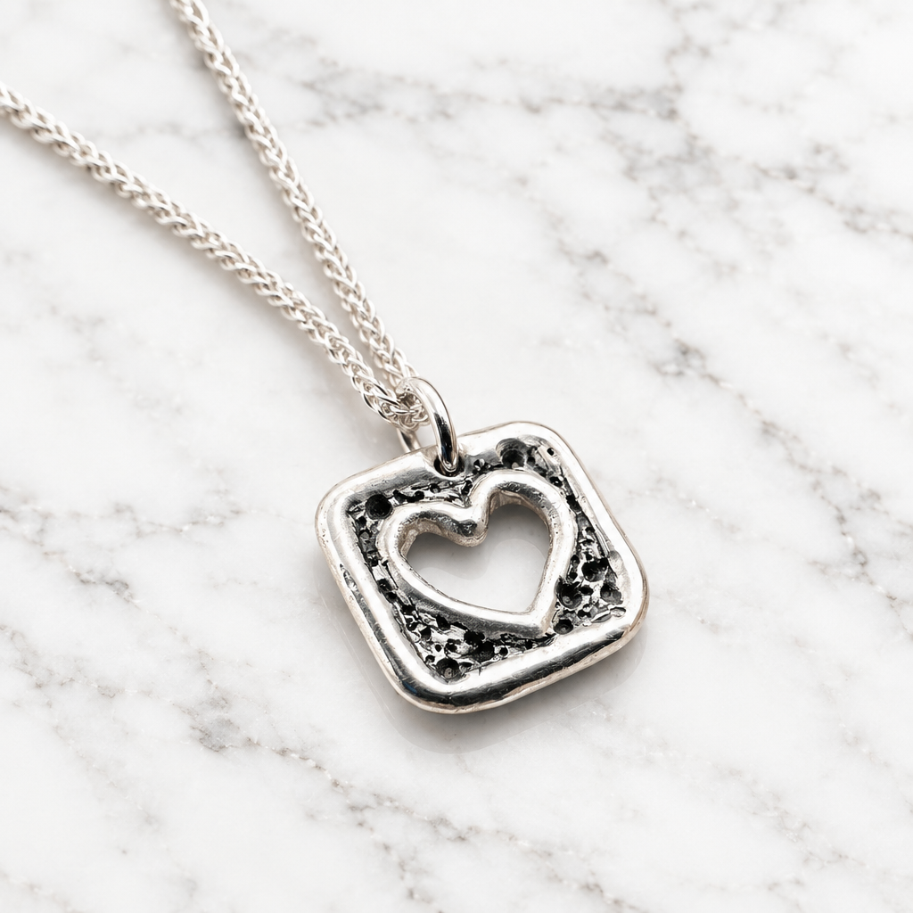 A silver pendant necklace featuring a textured, irregular square border with a hollow heart design in the center, suspended from a delicate silver chain against a clean white background.