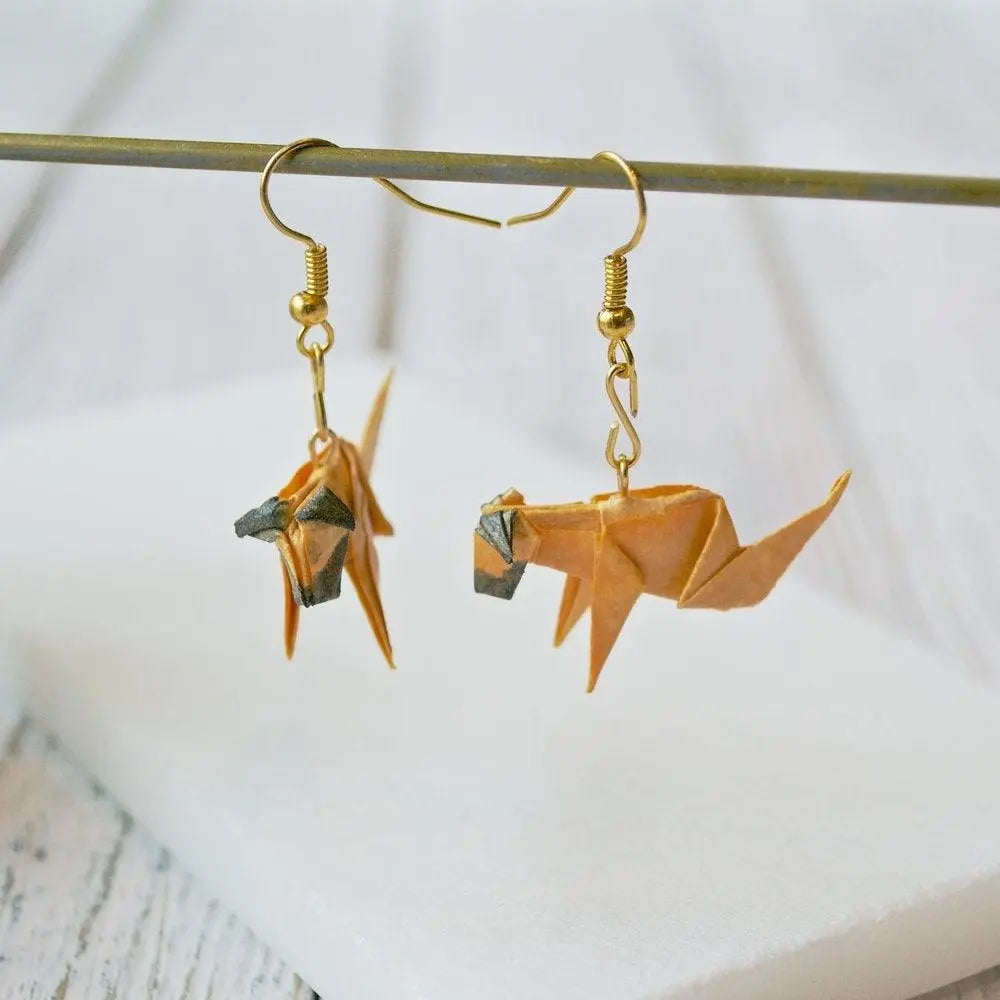 Pair of Origami Dog Paper Earrings hanging from a rod, showcasing their 3D folded dog design in tan and brown with gold hardware.