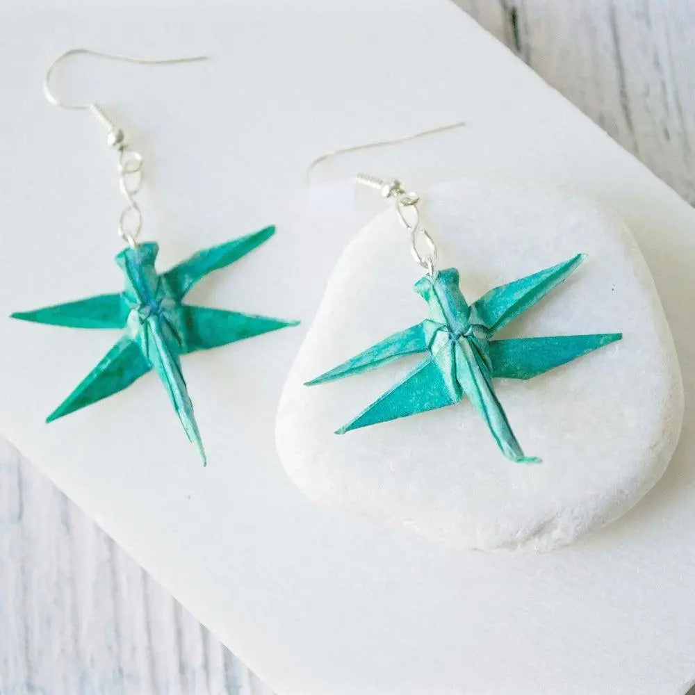 Paper origami earrings in a dragonfly design, handcrafted in teal paper with acrylic sealant, displayed on a white stone background.