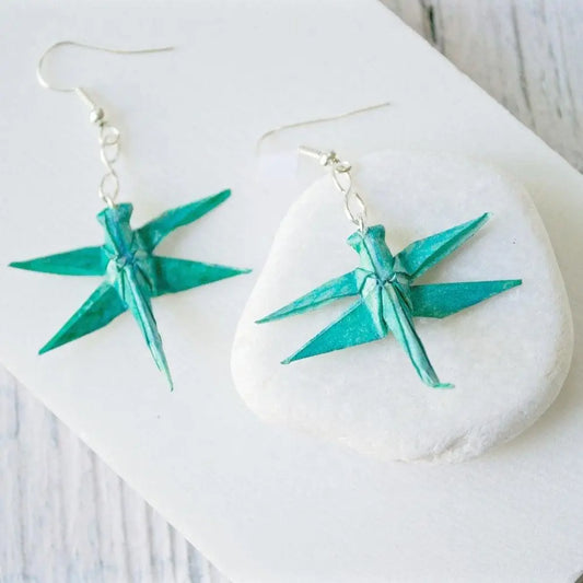 Paper origami earrings in a dragonfly design, handcrafted in teal paper with acrylic sealant, displayed on a white stone background.