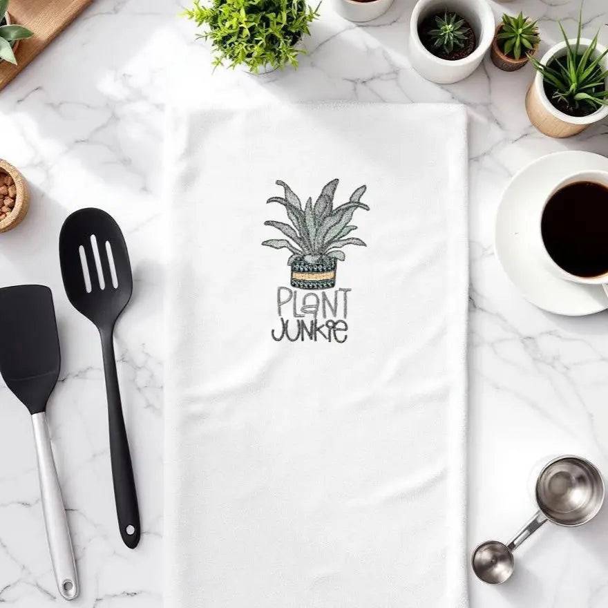Plant Junkie Embroidered Towel on marble kitchen counter with utensils, coffee, and potted plants. Absorbent fabric for stylish kitchen use.