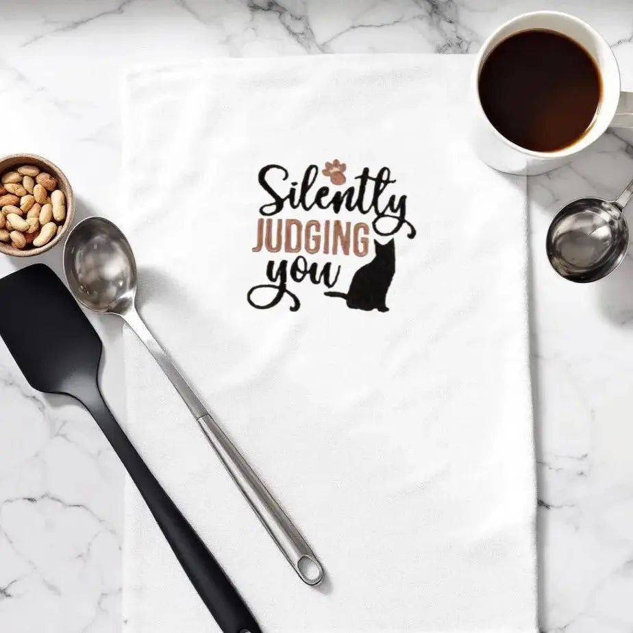 Silently Judging You Towel - Cat Embroidered | Kitchen Sass displayed on marble counter with coffee and utensils, ideal for quirky kitchen decor.