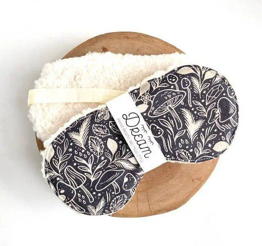 Cotton sleep mask for relaxation with black botanical print and soft Sherpa backing, displayed on a wooden surface. Ideal sleep aid accessory.