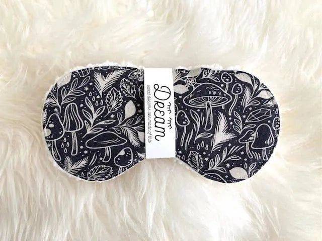 Cotton sleep mask for relaxation featuring black and white mushroom print, laid on a fluffy white surface. Soft Sherpa fabric for comfort.