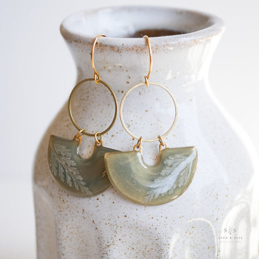 Fern Moon Shadow Earrings displayed on ceramic vase, featuring emerald green resin with fern imprint and gold hoop, perfect holiday accessory.