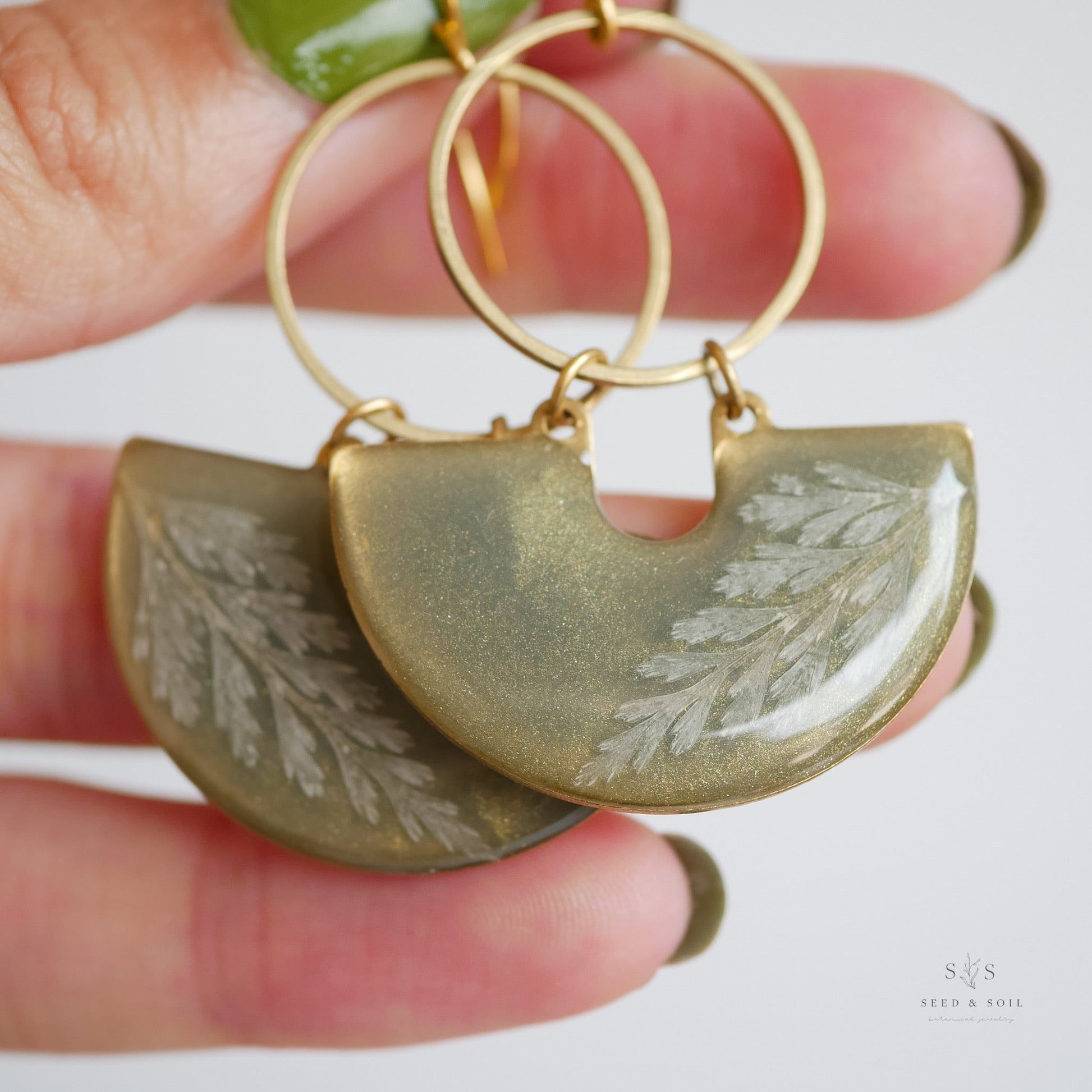 Hand holding Fern Moon Shadow Earrings with shimmering emerald resin and delicate fern imprint, gold hoop accent, holiday special edition.