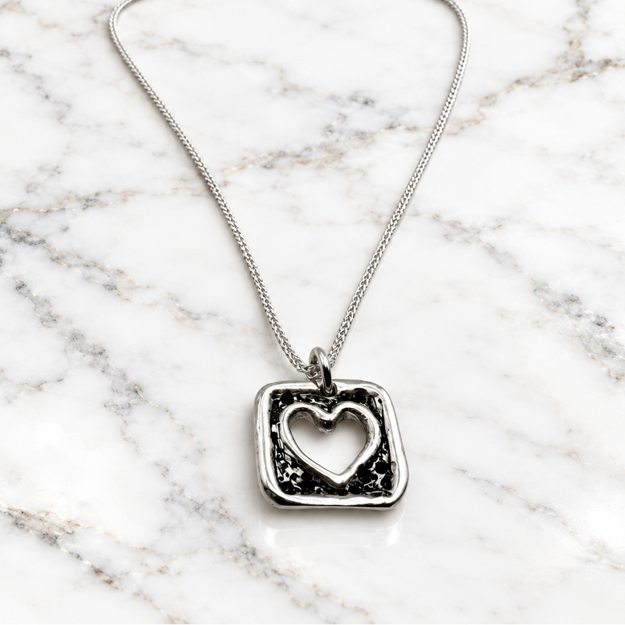 A silver necklace featuring a square pendant with a distressed blackened texture and a cutout heart in the center, suspended from a delicate silver chain against a plain white background.