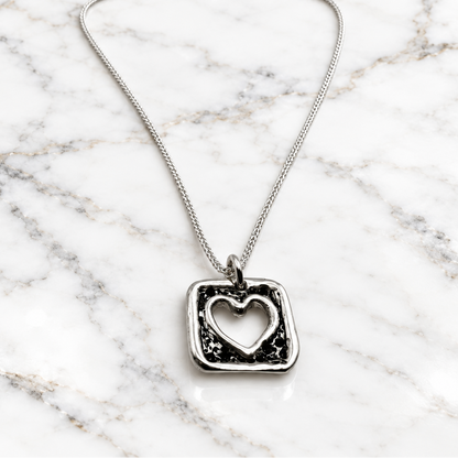 A silver necklace featuring a square pendant with a distressed blackened texture and a cutout heart in the center, suspended from a delicate silver chain against a plain white background.