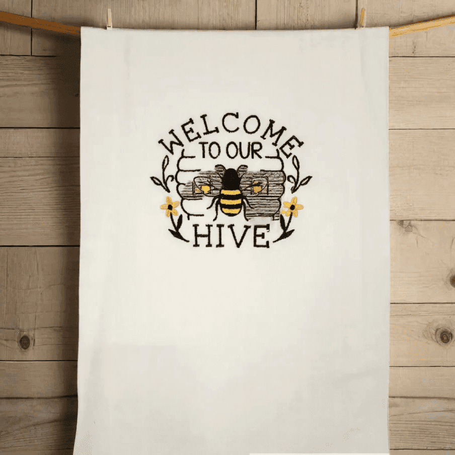 Bee embroidered towel hanging on a wooden wall, featuring 'Welcome to Our Hive' text and floral accents. Ideal for kitchen or bathroom decor.