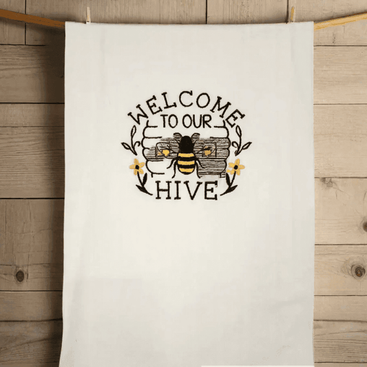 Bee embroidered towel hanging on a wooden wall, featuring 'Welcome to Our Hive' text and floral accents. Ideal for kitchen or bathroom decor.