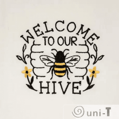 Close-up of bee embroidery with 'Welcome to Our Hive' text and yellow flowers on soft white fabric. Decorative and functional kitchen towel.