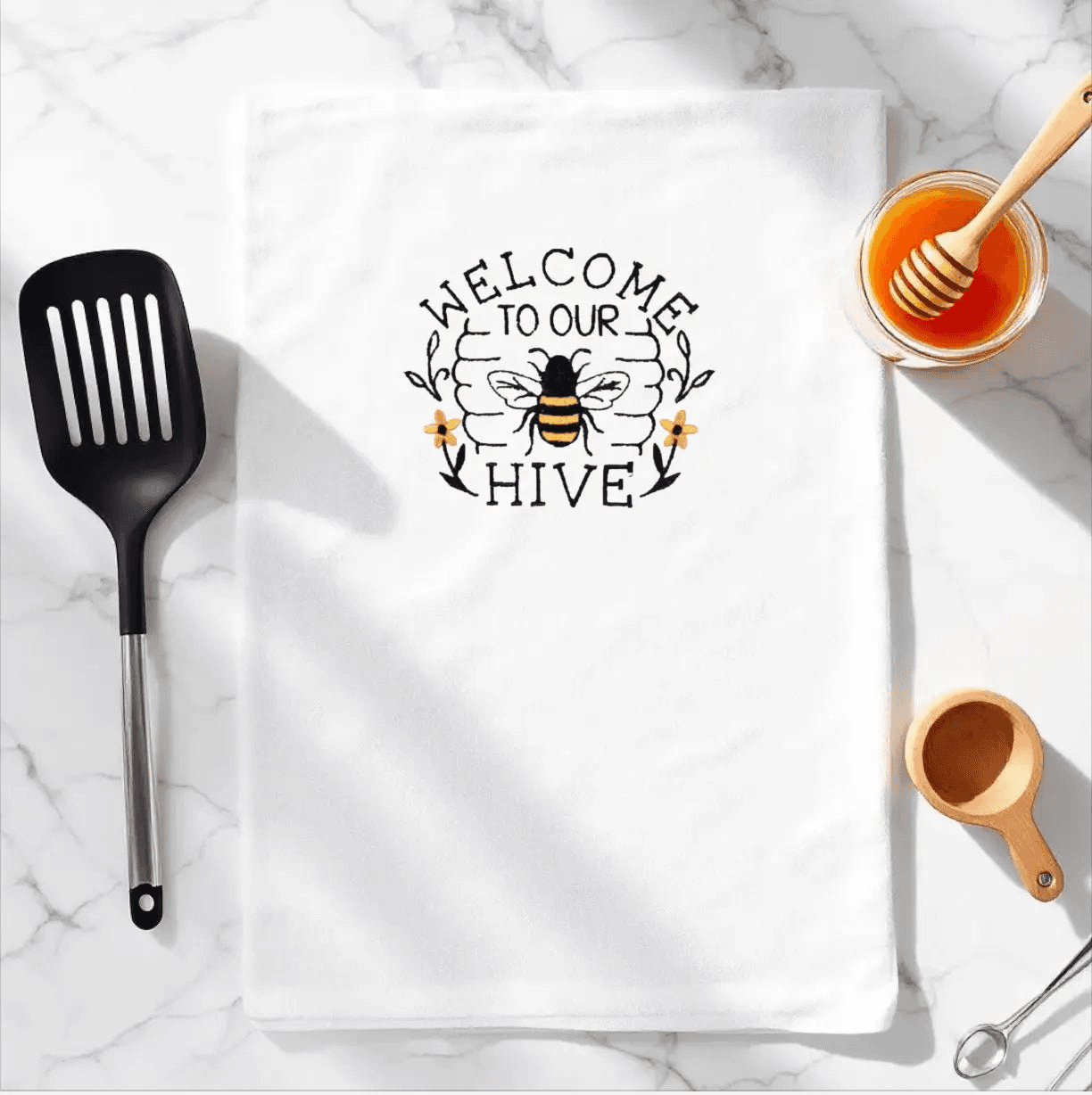 Embroidered towel with bee design and 'Welcome to Our Hive' text, displayed on a marble kitchen counter with honey and utensils. Soft and absorbent kitchen towel.