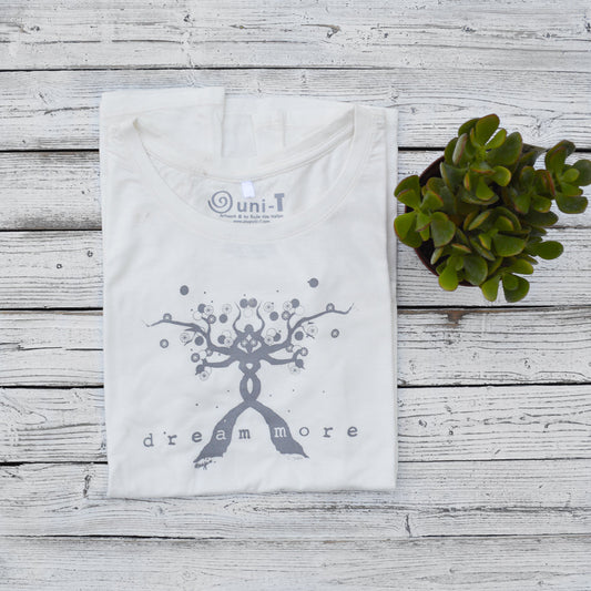 Folded white Women's Motivational T-Shirt in Bamboo & Organic Cotton with 'Dream More' tree design, laid flat beside a succulent.