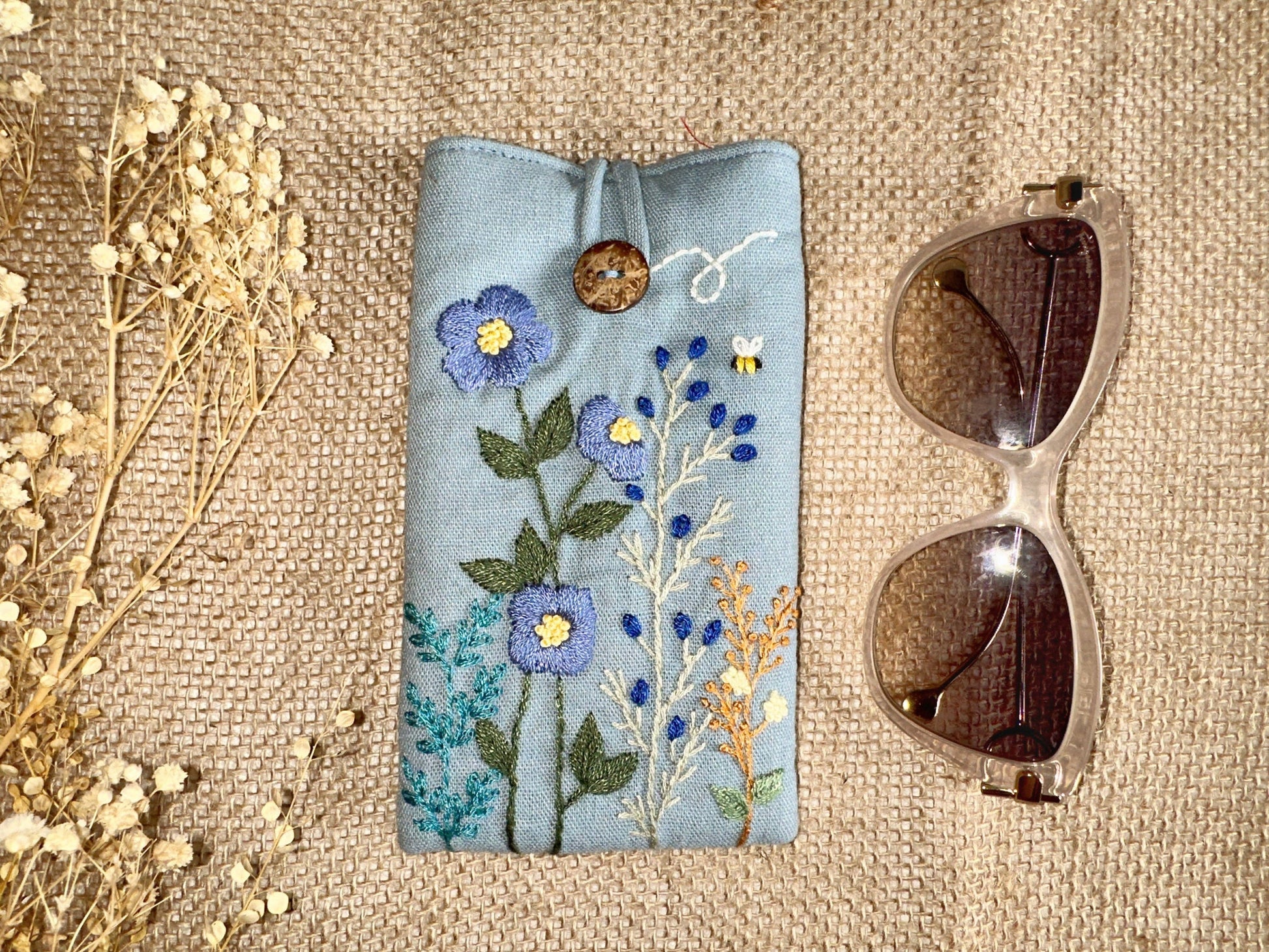 Blue embroidered linen glasses case with wildflowers and bee detail, featuring a wooden button and soft padded interior for protection.