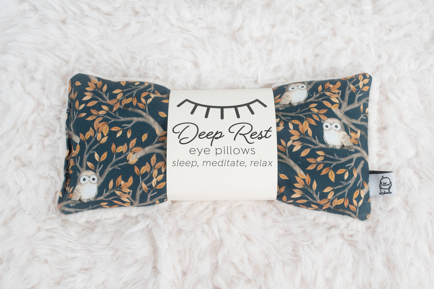 Weighted eye pillow for relaxation with owl and tree branch design, ideal for stress relief, meditation, and deep rest. Soft fabric on white fur surface.