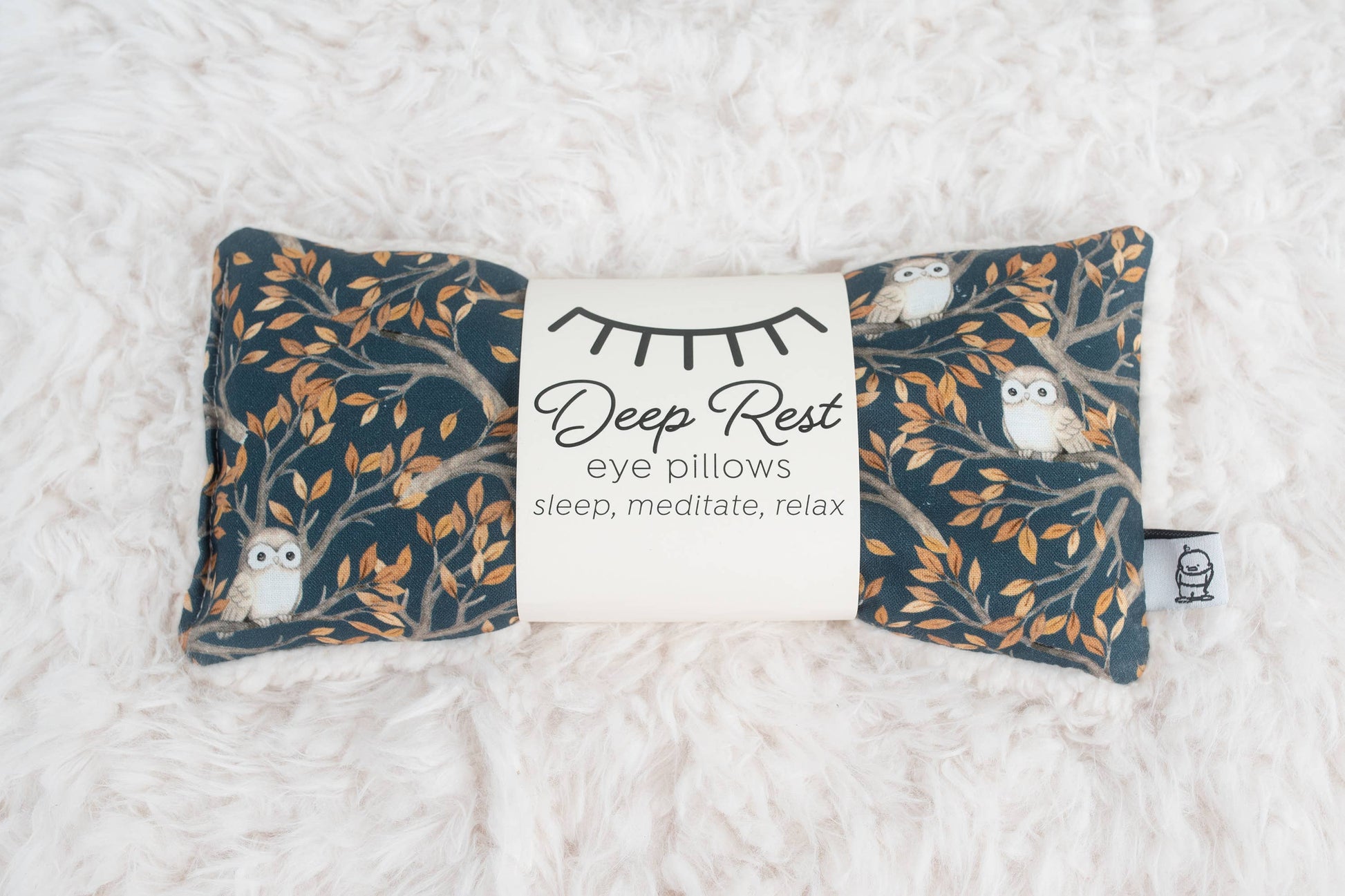 Weighted eye pillow for relaxation with owl and tree branch design, ideal for stress relief, meditation, and deep rest. Soft fabric on white fur surface.