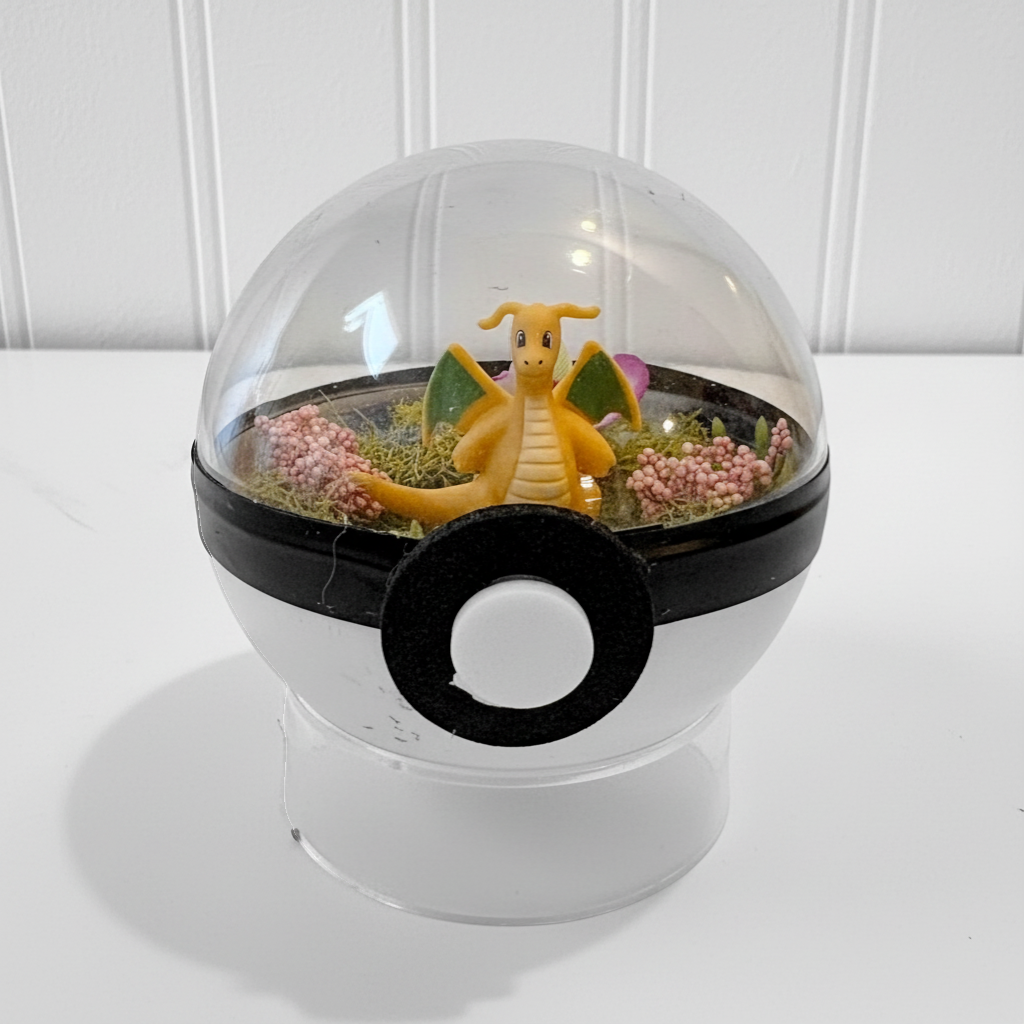 Uni-T Pokeball terrarium featuring a Dragonite figure inside a clear plastic sphere with moss and pink flowers