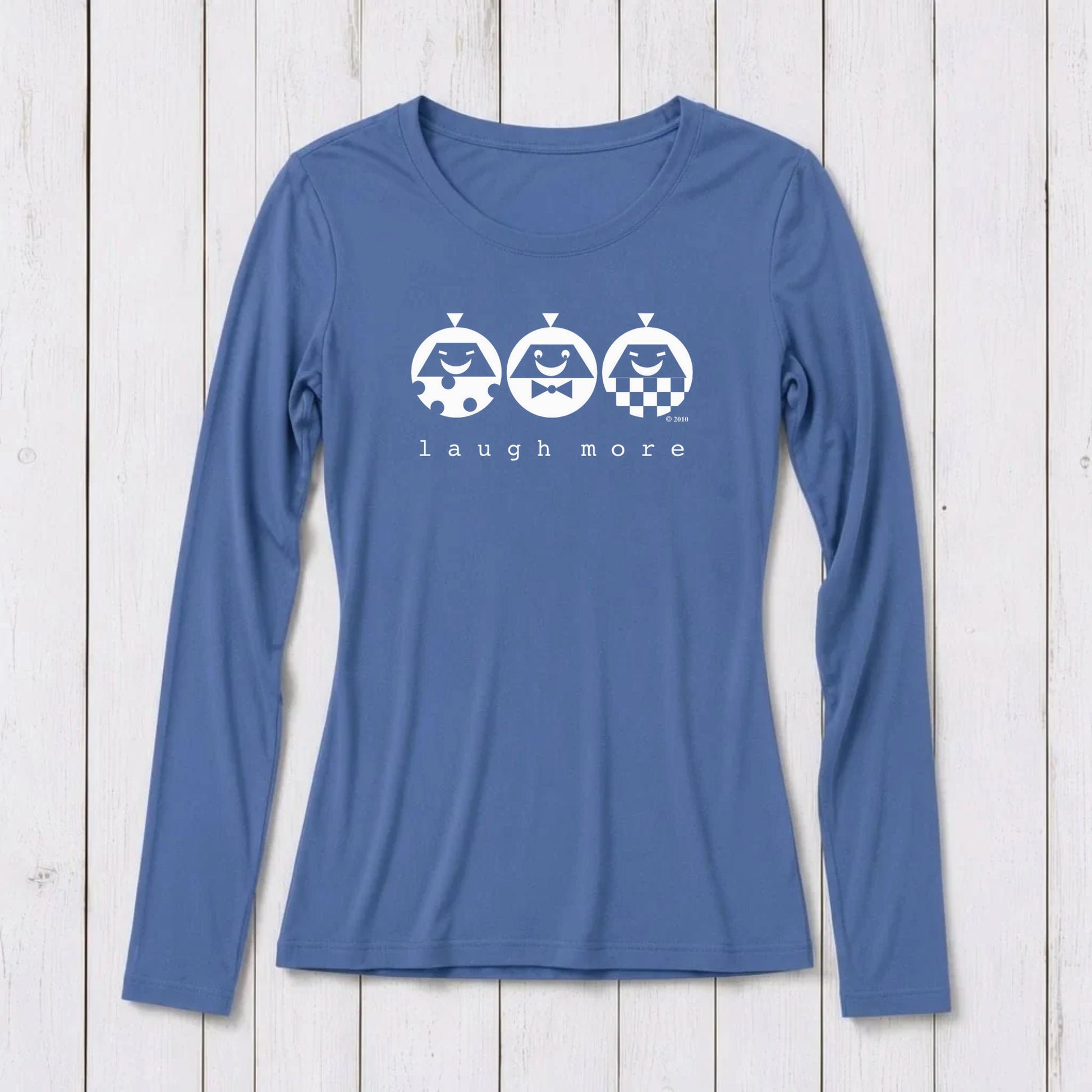 Blue long sleeve T-shirt with white graphic design featuring three smiling cartoon characters and the phrase 'laugh more' on a white wooden background