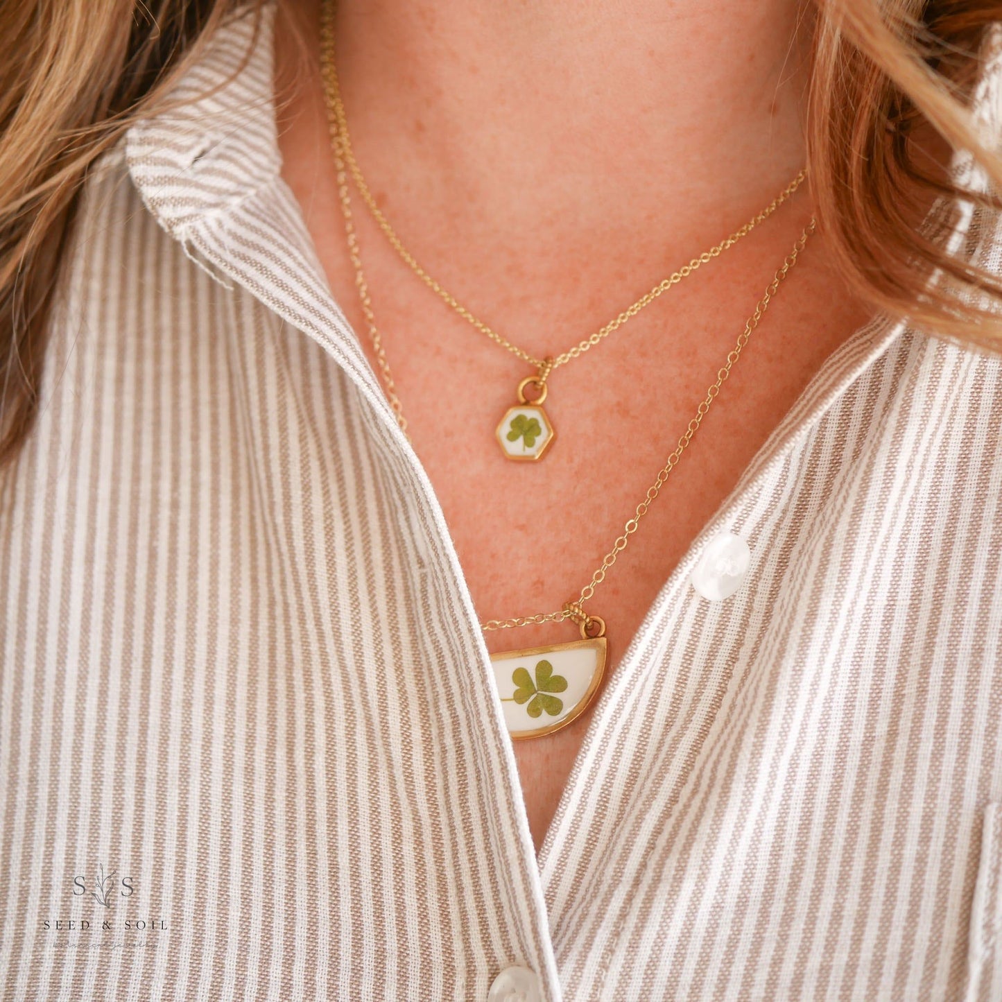 Layered clover pendant necklaces worn with a striped shirt. Features hexagon and half-moon styles in gold for a festive look.