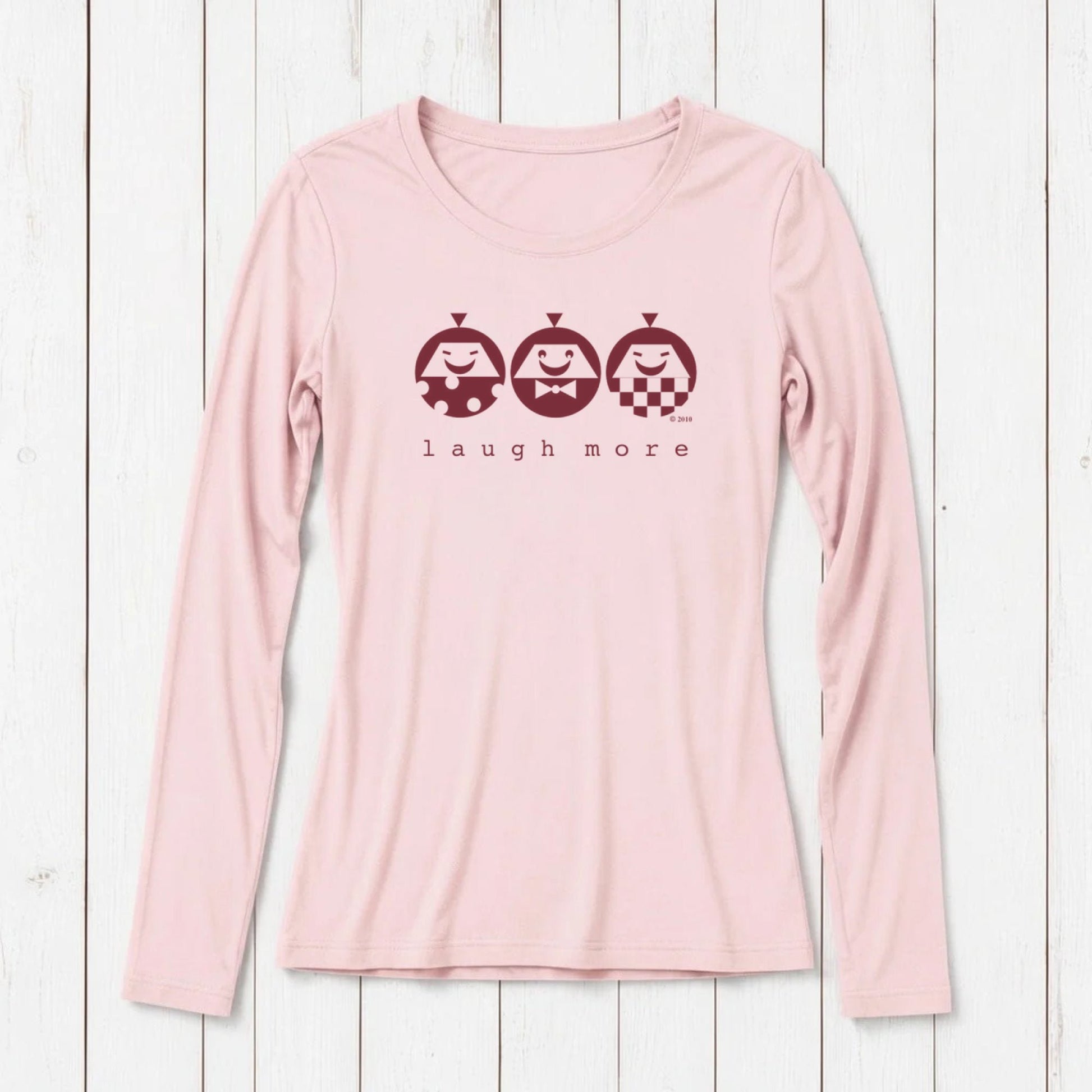 Light pink long sleeve T-shirt with a graphic design of three smiling cartoon characters and the phrase 'laugh more' in dark red, made from bamboo and organic cotton, laid on white wooden background