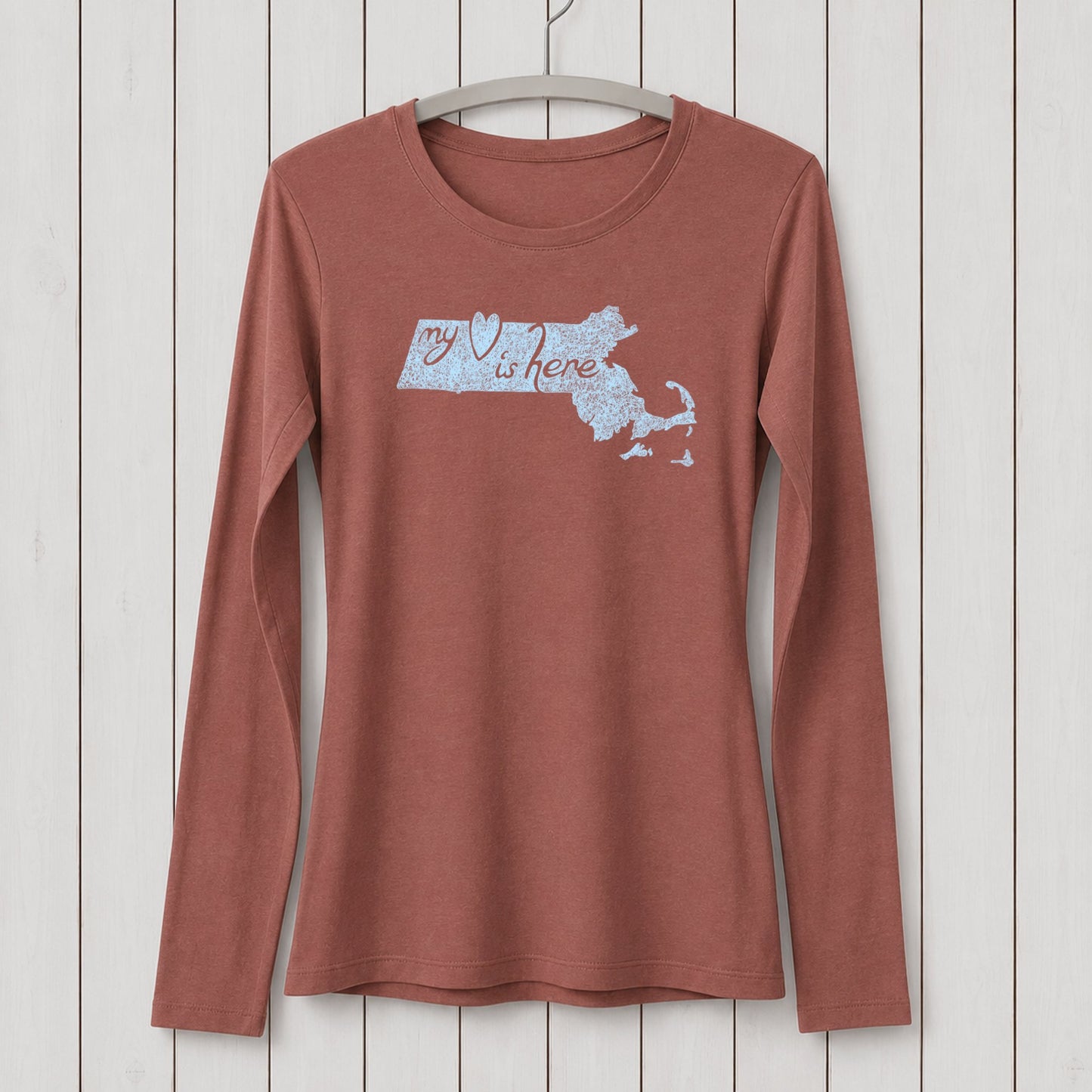 Rust-colored women's long sleeve T-shirt made of organic cotton and bamboo blend with a Massachusetts map and 'my heart is here' graphic design, hanging on a white wooden wall.