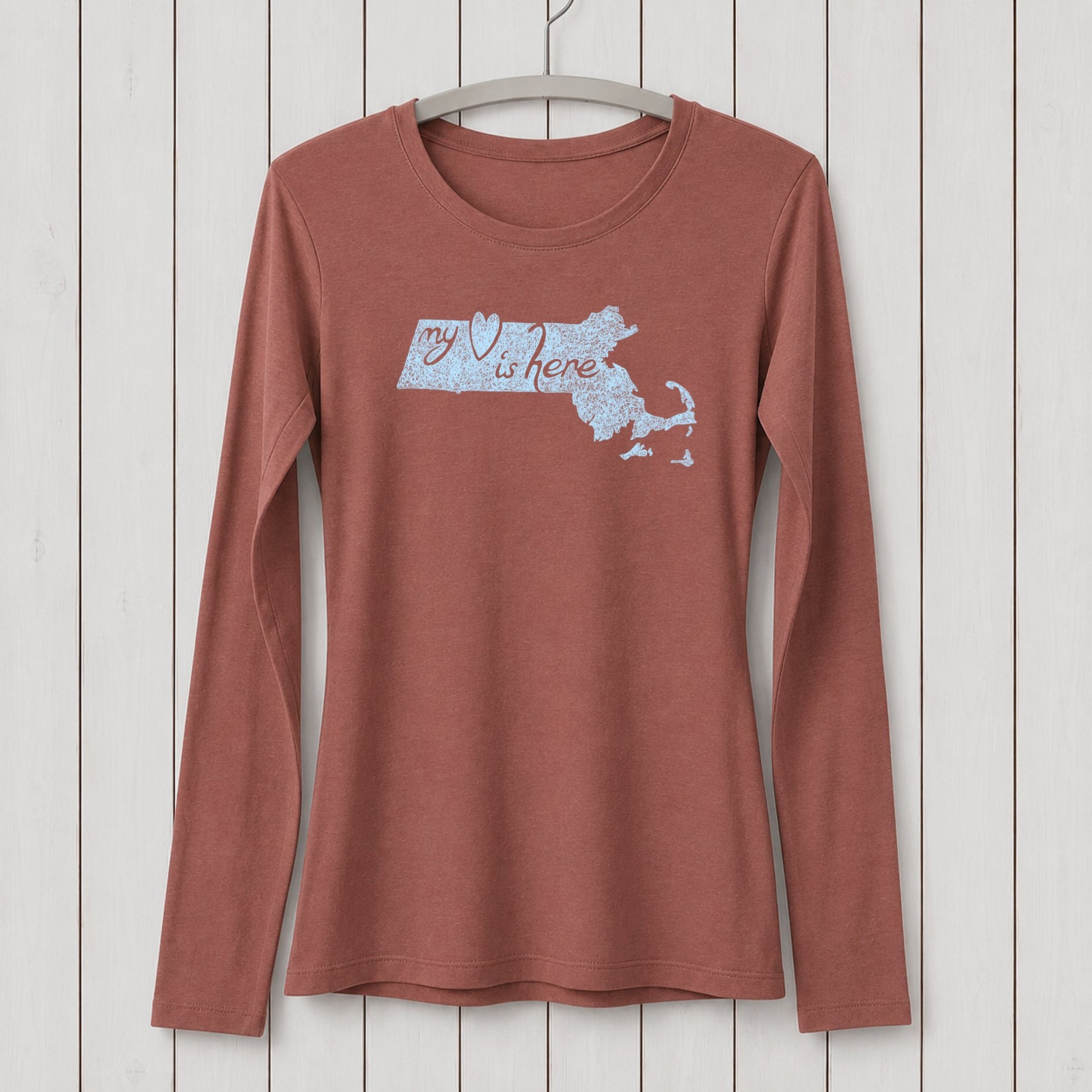 Rust-colored women's long sleeve T-shirt made of organic cotton and bamboo blend with a Massachusetts map and 'my heart is here' graphic design, hanging on a white wooden wall.