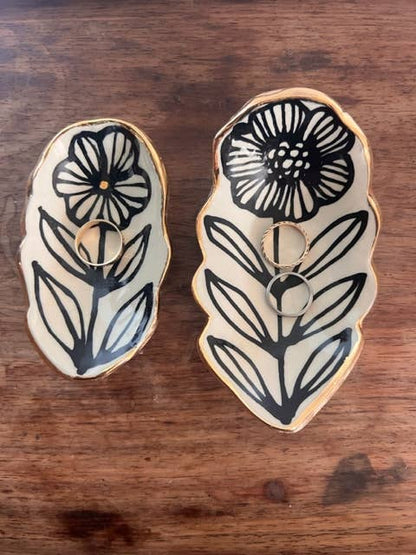 Two 5" handmade floral shaped 22K ceramic catchall jewelry dishes with black flower motifs and gold-rimmed edges, holding rings on wood surface.