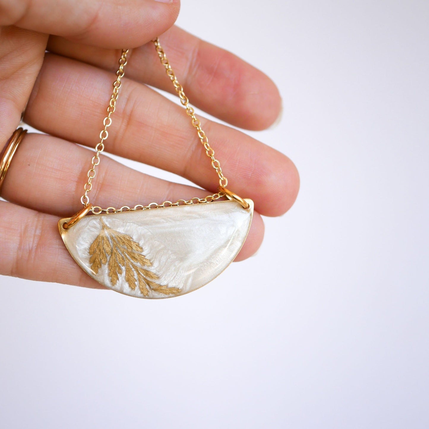 Hand holding the Classic Midas Collection Necklace showcasing its gold fern detail and half-moon resin pendant on a fine gold chain.
