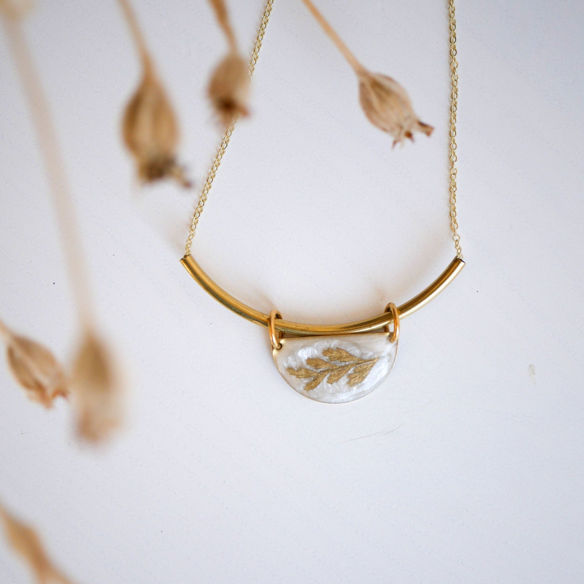 Shield-style Midas Collection Necklace with a curved gold bar and resin pendant featuring a golden fern, styled with dried floral accents.